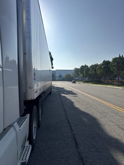 The image shows a semi-trailer truck parked on the side of a road in Fontana, California. The truck is white and has a reflective trailer. The road has yellow double lines, indicating a divided highway or a road where passing is prohibited. The background features a row of trees and some buildings, possibly warehouses, which are common in industrial areas. The sun is shining brightly, casting a long shadow from the truck across the road. A dark-colored sedan is visible in the distance on the road. The sky is clear and blue.