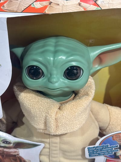 The image is a close-up, eye-level shot of a Grogu (Baby Yoda) toy. The toy is green with large, dark eyes and wears a tan robe. The toy is inside a yellow box, with the top edge of the box and a portion of the toy's packaging visible at the top. A label with text in multiple languages, including English, is visible in the lower right corner of the image. The background is a blurred yellow. The location in the city of Leers, France is not discernible from the image.