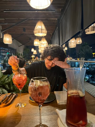The image displays a nighttime scene within a rooftop bar or restaurant, situated in Floridablanca, Colombia. The establishment features a ceiling composed of exposed wooden beams, from which numerous illuminated pendant lights with woven conical shades are suspended. A clear glass partition separates the interior from a panoramic view of a densely lit urban cityscape visible in the background.
In the foreground, a person with short, dark, curly hair, wearing a dark shirt, is seated at a wooden table. Their left hand operates a long stirrer within a large, stemmed glass containing a reddish beverage with ice and fruit pieces. Simultaneously, their right hand is positioned over their forehead, partially obscuring their right eye.
On the wooden table surface, to the left of the primary subject, another similar stemmed glass holds a reddish drink with ice and fruit. To the right, a clear glass pitcher or carafe is filled with an identical reddish liquid, fruit slices (including lemon), and ice. A small, dark, circular plate rests beneath one stemmed glass. A folded white napkin is placed beside the pitcher. A wooden serving board with cutlery is partially visible on the far left of the table. Several blurred figures of other patrons are discernible in the background, seated at distant tables.