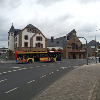 The media file captures an outdoor scene in Eisenach, Germany, featuring a city street with a bus and a train station. A bright yellow and red bus, advertising "Erste Hilfe Ausbildung" (First Aid Training) and "Hausnotruf" (Home Emergency Call), is prominently positioned on the right side of the road. The bus displays text in German, including phone numbers and a website address.

In the background, the Eisenach Hauptbahnhof (Main Train Station) stands with its distinctive stone architecture, featuring arched windows and a clock tower. A traffic light is visible, indicating a red signal. The sky is overcast, suggesting a cloudy day, possibly in the late morning or afternoon. The street is paved, with cobblestone sidewalks visible on the right. A pedestrian crossing is marked on the road.

Other elements in the scene include a traffic sign for a pedestrian crossing, streetlights, and a few cars and pedestrians in the distance. The overall impression is of a functional urban environment with public transportation and services being highlighted. The presence of the train station and bus suggests a travel hub or a transit point within the city.