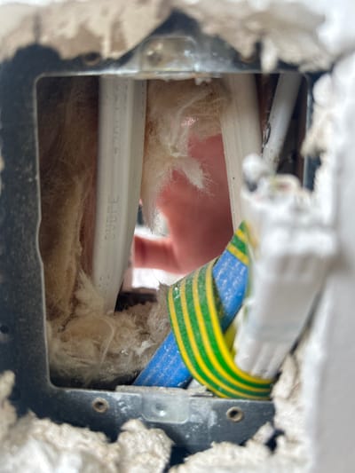 A square hole in a wall reveals wiring and insulation. White and gray plaster surrounds the opening, which is framed by a dark gray metal structure. Inside, white insulation obscures the view. Two white electrical cables run horizontally. A blue wire and a green and yellow striped wire snake through the space. A finger is partially visible behind the wires and insulation.