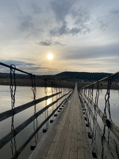 This image depicts a long, wooden suspension bridge stretching across a body of water. The bridge's deck is made of planks, and its sides are lined with wooden posts connected by twisted wire. The sun is low in the sky, creating a bright reflection on the water's surface. In the background, a forested area and some buildings can be seen. The location is Pepelyshi, Russia.