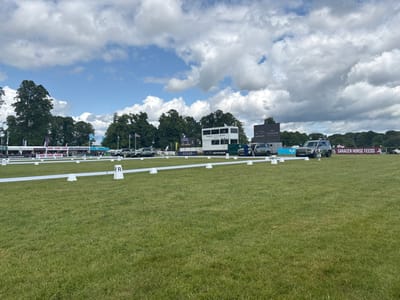The image shows a grassy field in Bramham, United Kingdom.  The field is surrounded by trees on the far side and has a line of white cones running across the middle.  There are three cars parked in the distance near the edge of the trees.  The middle car is a black Land Rover with a white roof. The cars are all facing the field and have an advertisement on the side.  The advertisement reads: "SARACEN HORSE FEEDS".  The cars are surrounded by people on the other side of the field. The image also shows a large white tent with an advertisement on the side. The tent is white with red lettering.  There are a few people standing by the tent and a single large screen in the background. The screen is behind the Land Rover with a message on it. There are also a few flags on the other side of the field. The sky is partly cloudy, the field is green and there is a small wooden structure in the background.  There is a large screen in the background with the word "BRAMHAM" on it.  The screen appears to be digital.  The image was taken from a low angle and looks toward the far side of the field.  The image was taken on a bright sunny day with many clouds in the sky.  The image appears to be taken during a sporting event.