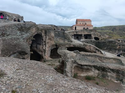 The image depicts the ancient cave city of Uplistsikhe in Georgia. In the foreground, large, weathered rock formations with visible cave entrances dominate the landscape. Patches of green vegetation sprout from the rocky ground. In the middle ground, several people are scattered across the rock formations, some sitting and others walking. A stone pathway winds through the area. Further in the background, a stone church with a red-tiled roof stands prominently on a hill. The sky is overcast with clouds.