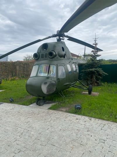 The image shows a military helicopter on display in Введенская Слобода, Russia. The helicopter, painted in olive drab, is positioned on a patch of grass next to a paved walkway constructed from light-colored stone blocks. The nose of the aircraft faces toward the viewer. A green fence is visible in the background, along with trees. The sky is overcast.