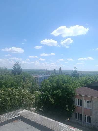 This is a wide-angle, high-angle shot of an urban landscape under a bright blue sky dotted with fluffy white clouds, likely taken in Florești, Moldova. In the foreground, the top of two corrugated metal roofs are visible, one with a flat grey surface and the other with ridged grey sheeting. A two-story building with a brown shingled roof and a facade of grey concrete blocks and reddish-pink panels occupies the right side of the frame. The building has multiple windows with white frames. Beyond the foreground structures, a dense canopy of lush green trees fills the mid-ground, obscuring much of the lower city. In the distance, several industrial smokestacks rise above the treeline, silhouetted against the horizon. The landscape appears somewhat undeveloped with rolling hills visible in the far distance. There are no people or animals visible in the image.