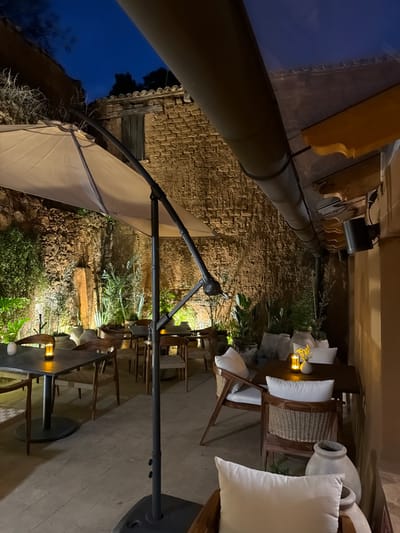 An outdoor dining area is illuminated by strategic lighting, highlighting stone walls and lush greenery. The setting features a long table with chairs, accented by white cushions and decorative elements, possibly candles or small lights. A large umbrella extends over part of the space, offering shade. The scene has a warm and inviting atmosphere, suggesting an evening setting with a combination of natural textures and ambient lighting.