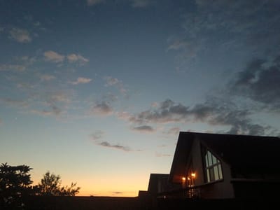 The image depicts a serene sunset scene with the silhouettes of buildings and trees against a gradient sky. The sky transitions from a soft orange at the horizon to a deep blue overhead, with scattered clouds catching the last rays of sunlight. Two modern-styled houses with prominent A-frame roofs are visible in the foreground, their windows reflecting the twilight sky. Lights are illuminated on one of the houses, casting a warm glow. To the left, the dark silhouettes of trees frame the lower portion of the image. The overall mood is peaceful and tranquil, suggestive of the end of a day. The time of day is clearly sunset. There is no visible text in the image. The location context suggests this scene is from Verkhnevesyoloye, Russia.