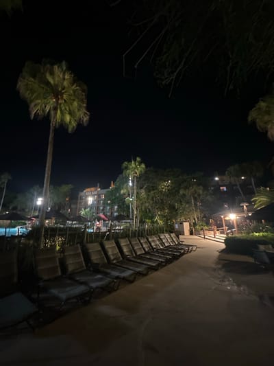 At night in Bay Lake, United States, a row of empty chairs sits near a path and pool. Palm trees are visible in the background, lit by ambient lighting. A building is seen further back. The area appears deserted, illuminated by artificial light sources.