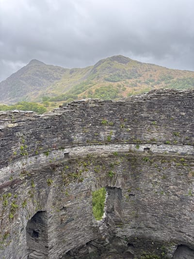 The image depicts a stone structure, possibly part of a derelict building or ancient ruin, situated in an outdoor environment in Llanberis, United Kingdom. The structure appears old, with weathered stones and visible signs of decay. Patches of vegetation, including grass and small plants, are growing on the stone surface. The structure has a hole or opening in its facade, revealing greenery inside. A mountain covered in vegetation rises in the background, partially obscured by overcast sky and fog. The setting suggests a historic site surrounded by natural landscape.