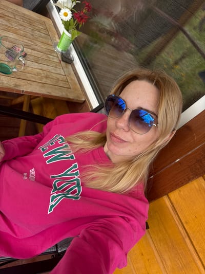 A woman with blonde hair and sunglasses is taking a selfie outdoors. She is wearing a pink sweatshirt with "NEW YORK" printed in green and white. She is sitting at a wooden table, where a glass mug with a green coaster and some items are visible. A vase with flowers sits on the edge of the table. The background shows a reflection of greenery and trees, suggesting a natural or semi-rural environment. The flooring is made of wooden planks. There is no indication of the location being Shuiskaya Chupa, Russia.