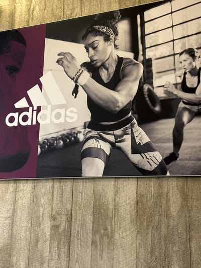 The image displays a large, vertical Adidas promotional poster mounted on an interior wall in Herzogenaurach, Germany. The poster occupies the upper two-thirds of the photograph. The left portion of the poster features a dark reddish-purple background with the white Adidas logo and text, partially obscuring the silhouette of a person's head and shoulder. The central and right portions of the poster depict a black-and-white scene of two women actively exercising in an indoor gym.

In the foreground, a dark-skinned woman with dark, curly hair tied back with a bandana is captured in a dynamic, bent-knee stance. She wears a dark sports bra, patterned athletic leggings, and multiple bracelets on her left wrist, with her arms bent and hands positioned forward. Her facial expression shows concentration. In the mid-ground to the right, a second, lighter-skinned woman is partially visible in a similar athletic pose, appearing to move forward. The gym environment behind them includes large, multi-paned windows, a large tire, and a row of kettlebells on the floor.

Below the poster, the bottom third of the photograph reveals a light-toned wooden floor with vertical planking. A narrow strip of a golden-toned textured surface is visible immediately above the poster's top edge.