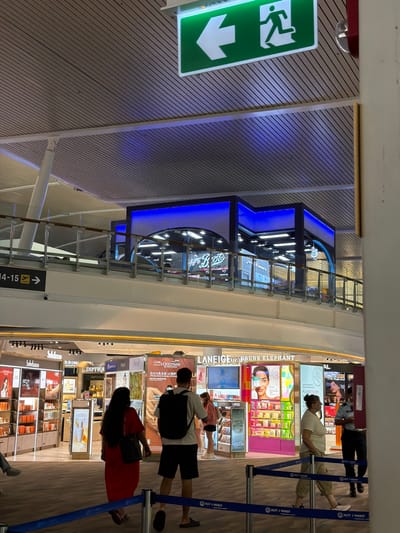 This image captures an indoor scene within what appears to be an airport terminal's retail area, likely a duty-free zone at Phuket International Airport. The perspective is slightly angled, looking across a shopping concourse.
Several people are visible, engaged in browsing or moving through the area. One person, wearing a light-colored top and light pants, stands near a waist-high blue barrier, looking towards the brightly lit product displays. Another individual in a red dress and carrying a dark handbag also observes the merchandise. Further back, a person in a uniform, possibly airport staff, is seen.
The setting features numerous illuminated display shelves showcasing a variety of cosmetic and beauty products. Brands prominently advertised include "LANEIGE," "DRUNK ELEPHANT," "L'OCCITANE EN PROVENCE," "DIPTYQUE," "Sulwhasoo," "le de peau BEAUTÉ," and "Fresh." Some displays feature models and promotional text like "PLEASE RESPONSIBLY" and "ACTHVE NDENS PROVEN POFORNICE." A Chinese phrase, "含20%乳木果 4X强护," is also visible on a L'Occitane display.
Architectural elements include a patterned ceiling or wall panel, bright overhead lighting, and a distinct structure with blue accent lighting on the left side of the frame. A prominent green emergency exit sign with a running figure and a downward arrow is visible on the left. A directional sign below the barrier indicates "14-15 ->," likely referring to gates. The blue barriers are repeatedly branded with "AOT / PHUKET," strongly indicating the location as Phuket Airport, which aligns with the provided location context of Mai Khao, Thailand. The overall impression is a bustling, well-lit commercial space.