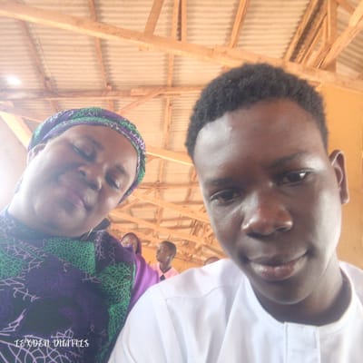 The image is a close-up selfie taken indoors, under a corrugated metal roof supported by wooden beams. In the foreground, on the left, a woman wearing a purple and green patterned headwrap and garment is looking slightly down. To her right and slightly in front, a young man wearing a white shirt is looking towards the camera. In the background, two other individuals, a young woman in a purple headwrap and a young man in a pink shirt, are visible. The lighting suggests it is daytime. The location is identified as Agbara, Nigeria.