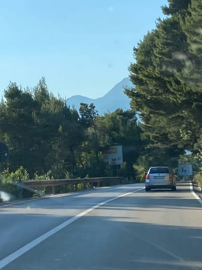 The image depicts a roadside scene near Blizikuće, Montenegro. A silver car is traveling on a paved road with a solid white lane marking. The road is bordered by lush green trees and vegetation. In the background, a distant mountain range is visible under a clear blue sky. A roadside sign is also present. The image appears to have been taken from within another vehicle.