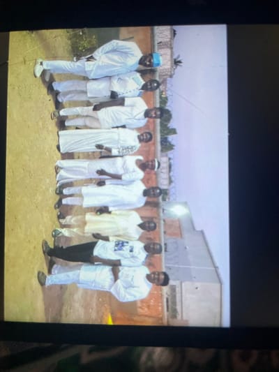 Nine adult males are depicted standing in a line, facing the observer, in an outdoor setting identified as the city of Mara, Nigeria. The majority of individuals are dressed in traditional light-colored, predominantly white, full-length garments or tunic-style outfits. One male wears a white graphic t-shirt with black trousers, and another wears a light yellow tunic over lighter trousers. A man positioned at the far left of the line wears a blue cap. The ground surface is unpaved reddish-brown soil. In the background, a light-colored wall topped with barbed wire or razor wire separates the immediate foreground from visible single-story buildings with flat roofs. Sparse green foliage is present to the left of the group. The scene is illuminated by natural daylight.