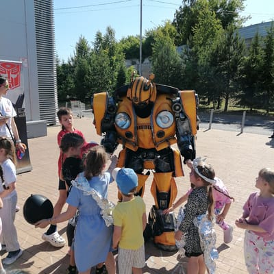 The image depicts an outdoor event in Трубачеевка, Russia, under clear, sunny skies. A person in a detailed yellow and dark gray Bumblebee costume, featuring prominent circular headlights on the chest and the Autobot logo, stands centrally, surrounded by several children. An adult female in a white t-shirt, wearing yellow-rimmed sunglasses on her head, is positioned on the left, observing the scene while holding a smartphone.
The children, of various ages, are actively engaging with the Bumblebee character. Some children are partially adorned with silver foil, suggesting a themed activity or costume element. One girl has pink face paint, and another in a blue dress holds a black spherical object. The ground is a light brown paved surface with visible chalk markings.
In the background to the left, a modern building with light gray vertical slatted panels features a large red Transformers Autobot logo and the Cyrillic text "ПГ" on a white banner. Further back, a line of dense green coniferous and deciduous trees separates the foreground from a visible section of an asphalt road with two light-colored bollards and a distant building with a gray roof. A stack of metal shopping carts is also visible behind the children near the building.