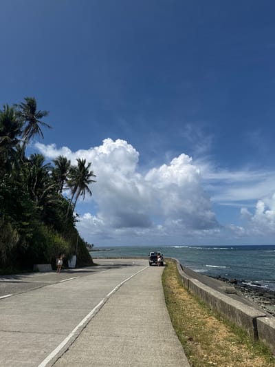 The image shows a road running along the coast in Burgos, Philippines. The sky is blue with large cumulus clouds. Palm trees grow on the left side of the road. A concrete barrier separates the road from the ocean on the right. A person in white clothing is standing on the side of the road. In the distance, a vehicle with two occupants is visible.