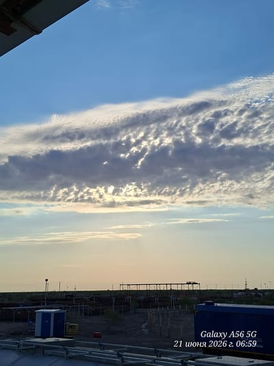 This is an outdoor, low-angle landscape photograph taken during the day. The sky occupies the upper two-thirds of the frame and features dramatic, layered clouds with sections of bright sunlight breaking through, particularly in the middle distance. The lower third of the image depicts an industrial or construction site under a soft, hazy sky. In the foreground, there are metal structures, possibly part of a walkway or railing. Further back, a blue utility box is visible, adjacent to a yellow cylindrical object and a red object. The middle ground reveals a landscape dotted with stacks of pipes or metal rods, and various industrial structures, including scaffolding and towers, suggesting an active worksite. The horizon is flat and features a line of distant trees or vegetation, with utility poles scattered across the landscape. The overall lighting suggests it is either early morning or late afternoon, with a warm, diffused light. The image also has text overlaid in the lower right corner, reading "Galaxy A56 5G 21 июня 2026 г. 06:59".