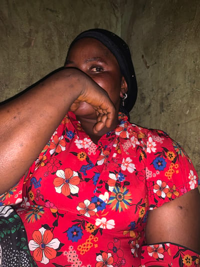 A person lies on a textured, earthy surface, their body angled towards the camera. They are wearing a vibrant red garment adorned with a colorful floral pattern of white, orange, blue, and yellow flowers. A black head covering is visible, framing their face. The person's right arm is raised, and their hand is positioned near their face, partially obscuring their features. One eye is visible, looking towards the camera, conveying a sense of weariness or introspection. The background is a rough, uneven surface, possibly dirt or stone, with some dark smudges. The lighting suggests an indoor setting or a shaded outdoor area, with no clear indication of the time of day or weather. The overall impression is one of quiet repose or perhaps discomfort. The context of "Project, Nigeria" provides a geographical and potentially thematic framework for the image. There is no visible text in the media file.