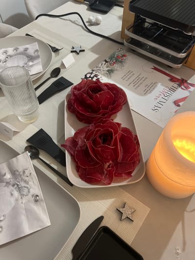 This media file captures a festive table setting prepared for a Christmas meal in an indoor setting. The scene is warmly illuminated, partly by a white ceramic candle holder emitting a soft, golden glow.
The main subjects are two plates of thinly sliced cured meat, artfully arranged to resemble red roses, indicating a sophisticated presentation for the meal. A "CHRISTMAS MENU" card is prominently displayed, featuring a red bow and winter-themed illustrations of pine cones and berries. The visible text on the menu details:
* **STARTERS**: "Chipe nature of paprika", "sive verte/mala" (likely "olives vertes/noires"), "flûte".
* **MAIN COURSES**: "Radette au fromage" (Raclette au fromage), "viande séchée" (dried meat), "cornichon et petit oignon" (gherkin and small onion).
* **DESSERTS**: "Gâteau au chocolat" (chocolate cake), "tarte en chocolat" (chocolate tart).
Tableware includes grey square plates, black cutlery (spoons and knives), and clear ribbed glasses set on light-colored woven placemats. Small black raclette pans are visible, along with a raclette grill at the top left, confirming the "Raclette" main course. Decorative elements like sparkling star ornaments and small white and silver baubles are scattered on the table, enhancing the holiday ambiance. The overall impression is of a cozy and meticulously prepared Christmas gathering, likely an evening event due to the candle lighting.
