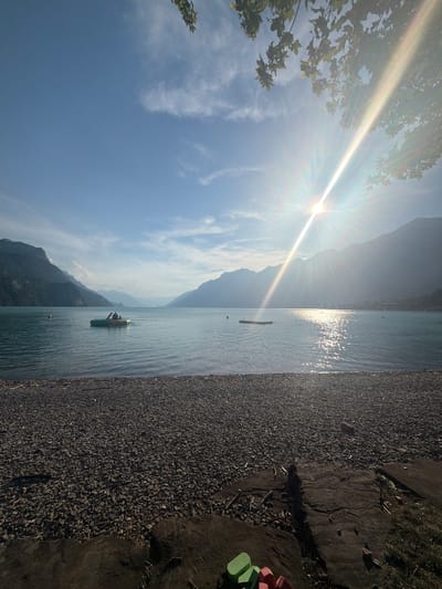 The image depicts a serene daytime scene at a lakeside beach in Brienz, Switzerland. The foreground is occupied by a dark grey pebble beach, with large rocks and some dry vegetation scattered across it. In the lower right corner, brightly colored children's toys (green and red blocks) are visible. The midground features the calm, blue water of a lake. A small, inflatable boat with two figures onboard is positioned on the water, slightly left of center. A yellow buoy is also present in the lake to the left of the boat. A wooden floating platform is located in the water to the right of the boat. The background is dominated by majestic, hazy mountains under a clear blue sky with scattered wispy clouds. The sun is bright and positioned high in the sky, casting strong rays and creating a glinting effect on the water. Tree branches with green leaves frame the upper right corner of the image.
