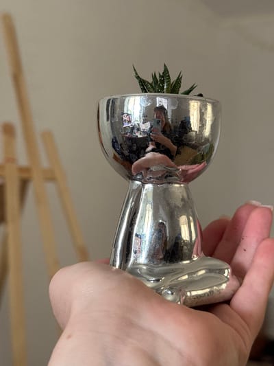 A person is holding a small, shiny, silver decorative object shaped like a meditating figure. The object has a small succulent plant in its bowl-like head. The figure is reflected in the shiny surface, along with the person holding it, who appears to be taking a selfie with a phone. In the background, a wooden easel is partially visible, suggesting the object might be on display or part of an art setup. The environment appears to be indoors with a neutral-colored wall.