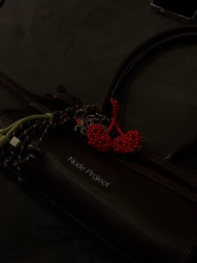 A dark, low-light image shows a partially visible brown leather handbag with silver lettering reading "Nude Project." Dangling from the handle is a red, beaded cherry keychain, and a knotted tassel with olive green, brown, and white elements. The bag sits on a dark, textured surface. The location is not discernible from the image.