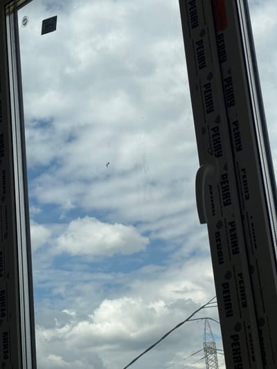 The image shows a close-up view of a window frame and the sky beyond. The window frame is made of white UPVC and has several labels on it. One label, near the top left, is circular with "AGC" written inside it. Another rectangular label below it has "25714" printed on it. Vertical labels with the word "PEHAY" are printed repeatedly along the frame. The sky is cloudy with patches of blue. In the distance, a power pylon is visible through the window, suggesting an outdoor or semi-outdoor setting within the city of Zhabkino, Russia. There are no people or animals in the image. The overall scene depicts a detail of a building's exterior.