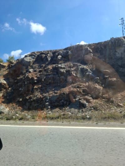The image depicts a view from inside a vehicle, likely moving along a paved road. To the left of the road is a rocky hillside or cliff face covered with vegetation. The sky is visible above the hillside, with some clouds present. An electrical transmission tower is seen at the top of the frame. The location is possibly near Ponor, Bulgaria.