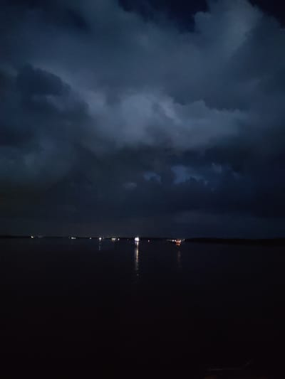 The image depicts a body of water at dusk or night, under a cloudy sky. In the distance, along the horizon, there are several lights, possibly from a settlement or vessels. The water is dark and reflects some of these lights. The sky is filled with dark, heavy clouds, suggesting potentially stormy weather. There are no visible people or specific structures that would indicate the exact location within the city of Каменники, Russia. The scene is characterized by low light conditions and a somber atmosphere.