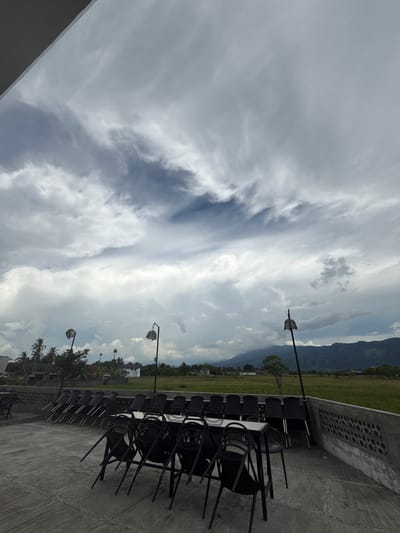 This image captures an outdoor seating area at what appears to be a restaurant or cafe. In the foreground, multiple black metal chairs are stacked and arranged around white tables on a concrete patio. Beyond the seating area, a low concrete wall with circular cutouts separates the patio from a lush green field that stretches into the distance. The field is bordered by a line of palm trees and scattered houses, suggesting a semi-rural or suburban setting. In the background, a mountain range with a hazy atmosphere looms under a dramatic, cloudy sky. Two decorative lamp posts with woven spherical shades are visible, one on the left and one on the right side of the frame. The overall impression is one of a quiet, possibly overcast day in a tropical location, specifically Meunasahmanyet, Indonesia. There are no people present in the image.
