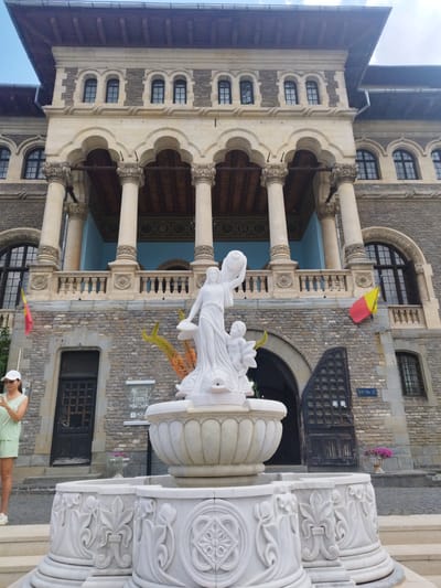 A full, eye-level shot captures the exterior of Cantacuzino Palace in Bușteni, Romania, a large, ornate building with classical architectural elements. The palace facade is constructed of stone, with a light-colored stone base and arched entrances. A prominent balcony with decorative columns and railings spans the lower section of the building. Several arched windows with dark frames are visible on the upper levels.
In the foreground, a white marble fountain features intricate carvings. A statue of a woman holding a drapery and a child is at the center of the fountain. The woman appears to be draped in a flowing gown, and the child is positioned near her feet.
To the left of the fountain, a woman wearing a light green outfit and a white cap is standing on a step, holding a smartphone in her hands and looking at it. She is looking down and to her right. A Romanian flag, colored red, yellow, and blue, is visible on a flagpole to the left of the fountain, near the building's entrance. To the right of the fountain, another Romanian flag is attached to the building.
The ground is paved with cobblestones and some stone steps lead up to the fountain. A few potted plants with pink flowers are visible near the building's entrance and to the right of the fountain. The overall atmosphere is one of grandeur and historical significance.