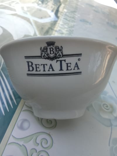 A white ceramic bowl with the black logo of "Beta Tea" is displayed on a table with a floral patterned tablecloth. The logo features a crown above the letter "B" enclosed in a shield, with the text "BETA TEA" below. The bowl is the primary subject, and the tablecloth provides a decorative background. The scene appears to be indoors, possibly in a home or cafe. There are no people or actions depicted. The lighting suggests it is daytime. The location is Sharg'un, Uzbekistan.