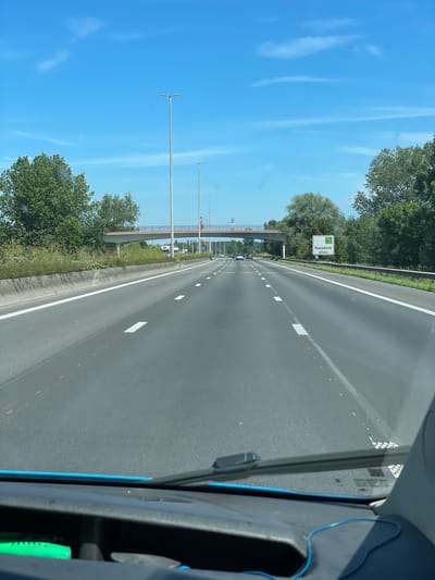 The image shows a view from the driver's seat of a vehicle traveling on a multi-lane highway. The highway has clear white lane markings and a concrete barrier on the left. Tall streetlights are visible along the side of the road. On the right side of the highway, a green and white road sign indicates "Haasdonk 400m" and "15a". A pedestrian bridge crosses the highway in the distance. Trees and vegetation line the sides of the highway. The sky is bright blue with some wispy clouds. There are other vehicles on the highway, including cars and trucks, all appearing to travel in the same direction. The foreground shows the vehicle's dashboard, windshield wipers, and a portion of the blue trim of the dashboard. The location is identified as being on the highway E17/A17 near Haasdonk, which is part of Temse, Belgium.