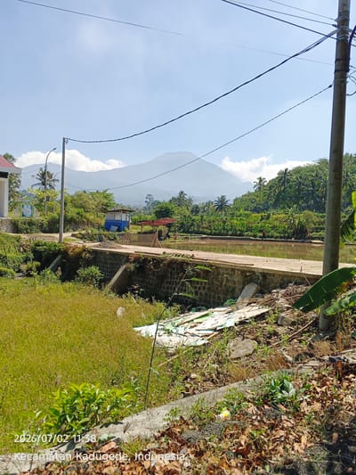 A daylight, eye-level, vertical-full shot captures a rural Indonesian scene in Kadugede. In the foreground, tall, dry grass covers the ground. A paved walkway with a stone retaining wall runs horizontally across the mid-ground. Beyond the walkway, a body of water, possibly a fish pond, reflects the sky. The background features a lush, green landscape with palm trees and dense vegetation, leading up to distant, hazy mountains under a bright blue sky with scattered clouds. A utility pole with electrical wires stands prominently on the right side of the image. Several small structures, including a blue and white building and a shelter with a red roof, are visible along the walkway and near the pond. Scattered debris, including broken wooden planks and white sheeting, lies on the ground in the lower right quadrant. A single individual in a red shirt is visible near the pond in the middle ground. The lower left corner of the image displays a timestamp and location: "2026/07/02 11:38 Kecamatan Kadugede, Indonesia."