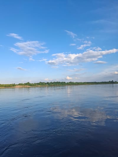 The image shows a wide, calm river with a distant shoreline covered in dense green trees. The sky is a vibrant blue, dotted with scattered white clouds. The water reflects the sky and the distant foliage. There are no people or objects visible in the scene. The environment appears to be natural and serene. The location is identified as Vvedenovka, Russia.
