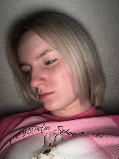 A close-up, tilted portrait shows a young person, likely a woman or girl, with blonde or light brown hair, resting indoors in Ligolambi, Russia. Her head is tilted to the right, with her face looking downwards and to the left. Her eyes are open, and her expression appears neutral or slightly pensive. Visible on her skin are some small blemishes and areas of redness on her cheek and forehead.
She is wearing a pink garment, possibly a hoodie or blanket, made of a soft, ribbed fabric. The garment features visible black text that reads "ecystudio School," though the initial part "ecy" might be "Lecy" or "Secy" due to partial obstruction. A faint, light-colored design, possibly depicting a cartoon animal like a bunny or cat, is also visible on the garment's right side. The background is a grey, textured wall or surface. The lighting is soft and ambient, characteristic of an indoor setting.