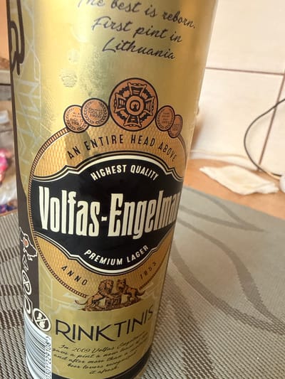 This is a close-up, angled shot of a gold-colored beer bottle. The bottle has a black and gold label with prominent text. The text "Volfas-Engelma" is the largest and most central piece of text on the label, written in bold, white, sans-serif font. Above this, "HIGHEST QUALITY PREMIUM LAGER" is written in a smaller, curved font. To the left of the main text, there's an inscription that reads "The best is reborn. First pint in Lithuania." There are also circular medals with text like "KAUNAS GOLD 1926" and "KLAI PEDA GOLD 1927" as well as "PARIS GRAND PRIX 1893". On the right side of the label, the text "RINKTINIS" is visible, and below it, a barcode and some smaller text, including a date that appears to be "2009" and mentions "beer lovers." The background is blurred, showing what appears to be a patterned surface, possibly a table or fabric, and a hint of light-colored walls or furniture. The lighting suggests an indoor setting, possibly during the day.
