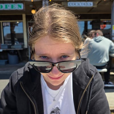 A headshot of a young person with blonde hair and blue eyes is centered in the frame, looking down at the camera with a slight smile. They are wearing black sunglasses and a white t-shirt with a silver necklace. A black zip-up hoodie is visible around their shoulders. In the background, there is a building with a sign that reads "OPEN" in green letters and another sign that says "COCKTAILS". Other people are blurred in the background. The scene appears to be outdoors, possibly a restaurant or bar, in Cannon Beach, Oregon, United States of America, indicated by the architecture and the style of the signage.