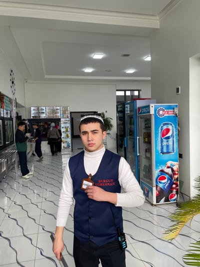 A young male stands in the foreground of an indoor cafeteria or restaurant located in Chinabad, Uzbekistan. He has dark hair, a neutral expression, and wears a white turtleneck sweater, a dark blue sleeveless vest, and dark pants. On his head, he wears a white and black traditional cap. A brown, shield-shaped nametag, bearing "BURGUT OSHXONASI" in red and "MAHMUD" in white, is pinned to his vest. A black and blue walkie-talkie is clipped to his right hip. His left hand holds a metallic object, possibly tongs or a serving utensil.
The background features a brightly lit, clean environment with white walls and ceiling. The ceiling is fitted with square recessed LED lights. The floor is tiled in white with a repeating dark blue wavy line pattern. To the left, a serving line with metallic counters and display cases is visible, where several individuals, including men and women, are present, appearing to select food or wait. A circular clock with a black face and white markings is mounted on the left wall. To the right, two upright Pepsi-branded display refrigerators contain various bottled and canned beverages. Text on one refrigerator includes "A'LO SERV" and other Uzbek script. A dark green leafy plant is partially visible in the bottom right corner.