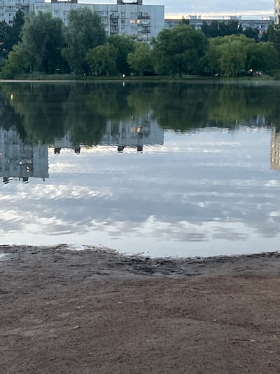 This media file is a vertical, close-up shot of a body of water reflecting buildings and trees. The scene depicts a waterfront area with apartment buildings on the left, a line of trees, and the water. The right side of the image shows a textured, dark surface, possibly a bank or pathway.

The water's surface is rippled, creating a distorted reflection of the environment. In the reflection, we can see the geometric patterns of apartment buildings with windows, interspersed with the lush green shapes of trees. The sky is reflected in the water, appearing as a mix of light and dark, cloudy patches. A few lights are visible in the windows of the buildings, suggesting it might be dusk or early morning.

The overall impression is one of a calm, urban or suburban landscape. The focus on the water's reflection adds an artistic and slightly abstract quality to the image. There is no visible text in the image. The location context is Vaganovo Massiv, Russia.