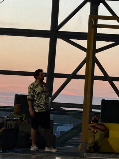 The image depicts a group of individuals in what appears to be an indoor waiting area, possibly an airport lounge or a similar public space, at sunset. The sky outside is visible through a large window, exhibiting hues of orange, pink, and pale yellow. Structural elements of the building, such as dark metal beams and a prominent yellow support column, are prominent in the foreground and midground.
In the center of the frame, a man with dark hair and glasses stands, wearing a short-sleeved, patterned shirt and dark shorts. He is looking to his right. To the left, another man, with a shaved head and wearing a green patterned shirt, is seated and appears to be holding a small object. Further to the right, a third man is reclining on a yellow surface, his eyes closed, wearing a dark red shirt and light-colored pants. Several dark seating structures are visible behind the standing man, and a dark suitcase is positioned near the seated man on the left. The overall atmosphere suggests a moment of rest or waiting. This scene is situated within Zaventem, Belgium, likely at Brussels Airport.