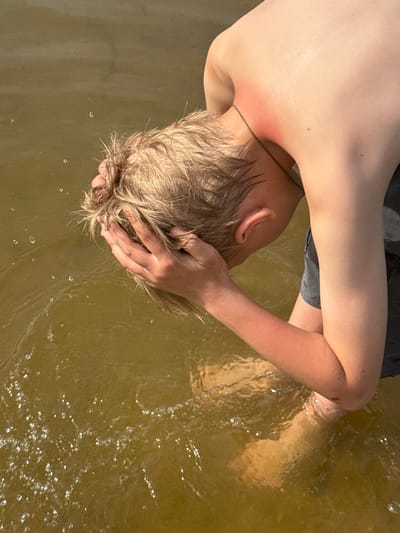 A boy is submerged in murky water, possibly in Novosinkovo, Russia. He has light blonde hair and is partially submerged. He rests his head in his hands, which are also in the water. He appears to have a sunburn on his back. His feet are also submerged in the water.