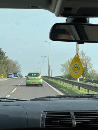 The scene is viewed from inside a vehicle, looking forward through the windshield on a road in Brentwood, United Kingdom. The vehicle's dark gray dashboard, windshield wipers, and two air vents are in the immediate foreground. A rearview mirror, positioned centrally at the top, reflects the upper portion of a person's head, revealing dark hair pulled back. Suspended from the rearview mirror is a yellow, teardrop-shaped air freshener, marked "COYENA" and featuring a blue-rimmed smiling sun graphic.

Ahead on the asphalt road, a light green Vauxhall Corsa, bearing the UK registration plate "EO59 KLF," travels in the same lane. Further ahead, two blue vehicles are visible in the left lane. The road features white lane markings. To the right, a metal guardrail parallels the road, with multiple utility poles or streetlights visible beyond it. Trees, some with bare branches and others evergreen, line both sides of the roadway. A partially obscured yellow circular road sign is present on the left verge. The sky overhead is clear and light blue.