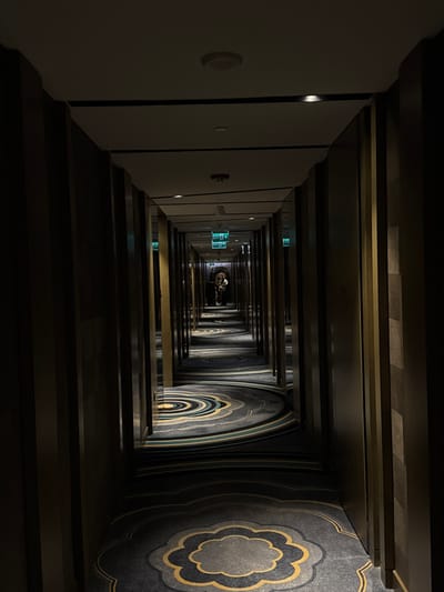 An enclosed hotel corridor creates a claustrophobic perspective. The left wall is lined with white doors, while the right features patterned carpet. Overhead, a coffered ceiling stretches into the distance. Diminishing perspective emphasizes the length of the corridor, culminating in a distant point where a figure is partially visible. Emergency exit signs are interspersed along the left wall. Overall, the composition conveys a sense of depth and confinement.