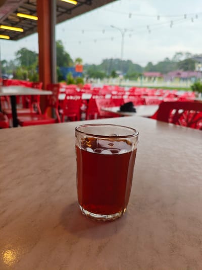 A close-up, eye-level shot focuses on a glass of dark amber liquid, possibly iced tea or coffee, situated on a light brown, speckled table. The glass, made of clear, textured glass, is half-filled with the beverage. The background is blurred but reveals an outdoor seating area with numerous bright red plastic chairs arranged around tables. The scene appears to be a food establishment, possibly a hawker stall or a restaurant, in Kota Tinggi, Malaysia, given the typical outdoor seating arrangements and bright red chairs commonly found in such locations. The lighting suggests it is daytime, with an overcast sky visible in the distance. The overall ambiance is casual and relaxed, hinting at a typical Malaysian roadside eatery.