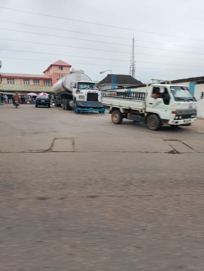 An overcast day is captured in Isheri, Nigeria, showing a street scene with vehicles and buildings. In the foreground, a white flatbed truck with a black metal rack is moving to the right, its driver visible inside. Behind it, a large white tanker truck, also moving to the right, is partially obscured. To the left of the tanker, a black sedan is parked under umbrellas, with a person on a motorcycle riding past it. In the background, a multi-story building with a red roof is visible, along with other structures and a distant telecommunications tower. The ground is paved with concrete, showing cracks and manhole covers. Power lines crisscross the sky.