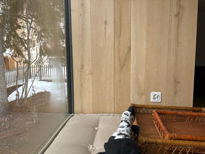 The image shows a person sitting on a cushioned bench, their feet resting on a wicker table. The person is wearing black pants and socks with a black and white design. The bench is attached to a wall with a large window and wooden panels. The wall has a white electrical outlet. The window offers a view of a snowy outdoor area with trees and a wooden fence. The coordinates 46.514, 9.859 place this scene in Celerina/Schlarigna, Switzerland.