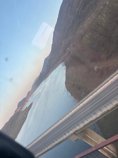 The image presents an elevated, panoramic view of a landscape identified as being in the city of Struya, Bulgaria. The composition is diagonally oriented from a rotated perspective.

In the midground, a concrete bridge spans across a wide body of water. The bridge features an asphalt road surface with two distinct lanes, marked by a solid white line on the viewer's left and a dashed white line on the viewer's right. Metal guardrails are present on both edges of the bridge deck. No vehicles or individuals are visible on the bridge.

The body of water, likely a lake or reservoir, occupies a significant portion of the scene, stretching from the left background towards the right midground, underneath the bridge. Its surface is calm and reflects the sky, appearing light blue-grey. Along the distant left bank of the water, a faint cluster of lights or structures suggests a settlement.

The background is dominated by a large mountain range, heavily covered in bare deciduous trees, indicating a late autumn or winter season. These mountains recede into the distance on the left. The sky, visible in the upper left, displays a gradient from clear blue at the top to a pale orange-yellow hue near the horizon, characteristic of twilight conditions. A faint white aircraft contrail is discernible in the upper sky.

The immediate foreground, particularly the bottom right, includes dark, indistinct structural elements and a red-brown bar, consistent with a window frame or viewing aperture. Several reflections, including a prominent rectangular shape and circular smudges, are visible on the transparent surface through which the photograph was captured. No people or specific actions are depicted within the scene.